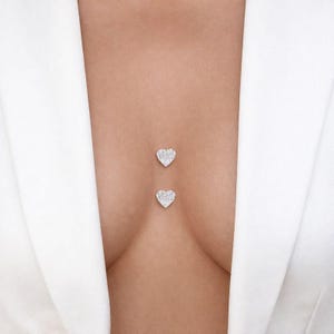 May include: Two heart-shaped body jewellery pieces, each encrusted with small, sparkling stones. The jewellery is positioned vertically on the body. The background is a white blazer.