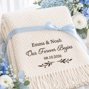 Könnte beinhalten: Eine cremefarbene Strickdecke mit Fransen, umwickelt mit einem hellblauen Satinband. Die Decke ist mit dunkelbraunem Text bedruckt, der Emma & Noah, Our Forever Begins, 08.10.2026 lautet, sowie einer dekorativen Zweiggrafik.