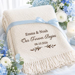 Peut inclure: Un plaid tricoté de couleur crème entouré d'un ruban de satin bleu clair. La couverture est brodée avec le texte Emma & Noah, Our Forever Begins, 08.10.2026, accompagné d'un petit motif graphique de branche en brun foncé.