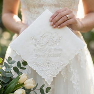 Peut inclure: Un mouchoir blanc avec des bords festonn&eacute;s, brod&eacute; de la lettre "B", d'&eacute;l&eacute;ments floraux et des noms "Anthony & Anita" ainsi que la date "25 mars 2026". Le mouchoir est tenu par une personne portant une robe de mari&eacute;e.