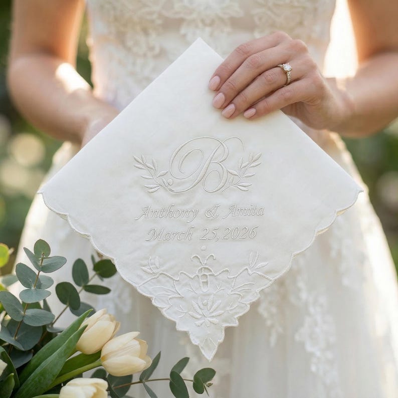 Peut inclure: Un mouchoir blanc avec des bords festonn&eacute;s, brod&eacute; de la lettre "B", d'&eacute;l&eacute;ments floraux et des noms "Anthony & Anita" ainsi que la date "25 mars 2026". Le mouchoir est tenu par une personne portant une robe de mari&eacute;e.