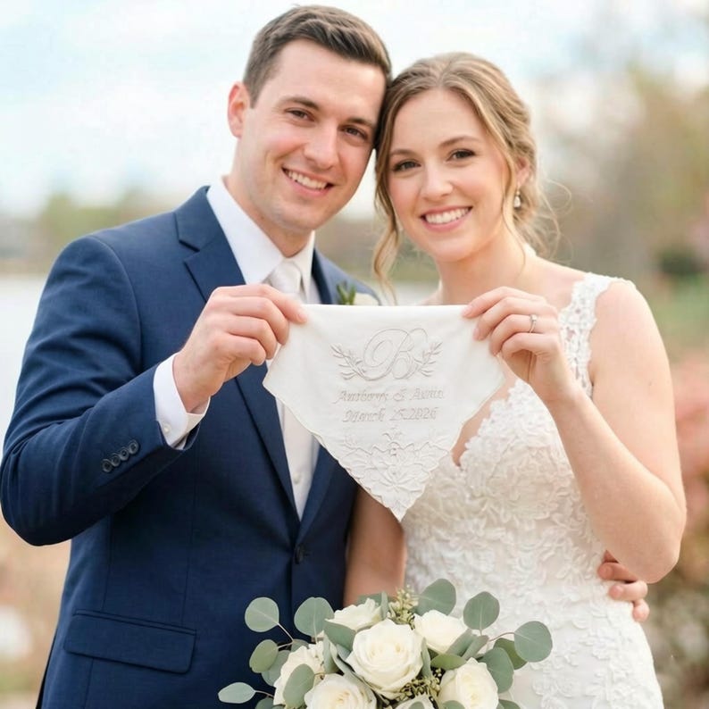 Peut inclure: Un couple souriant tient un mouchoir blanc avec des initiales brod&eacute;es et une date de mariage. Le mouchoir a une bordure en dentelle d&eacute;licate et des motifs floraux. Le mari&eacute; porte un costume bleu marine et la mari&eacute;e une robe de mari&eacute;e en dentelle blanche. Un bouquet de roses blanches est au premier plan.