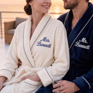 May include: A cream-colored plush robe with grey piping and the embroidered text Mrs. Bennett in navy blue, alongside a navy blue plush robe with white piping and the embroidered text Mr. Bennett in white, both featuring long sleeves and belts.