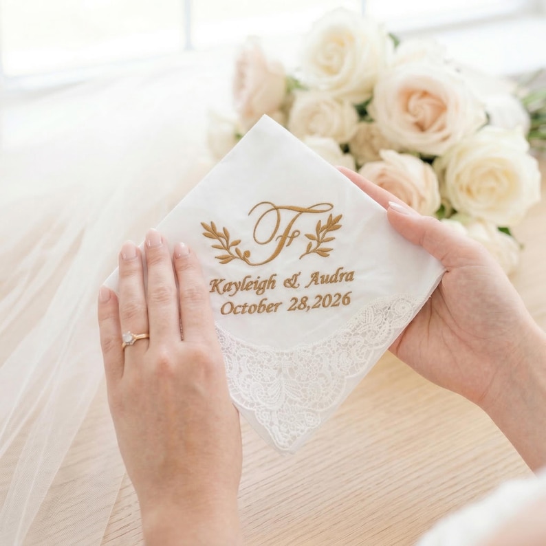 Peut inclure: Mouchoir blanc avec broderie dor&eacute;e, avec les initiales "F" et un motif floral. Le texte "Kayleigh & Audra October 28, 2026" est &eacute;galement brod&eacute;. Le mouchoir a une d&eacute;licate bordure en dentelle.