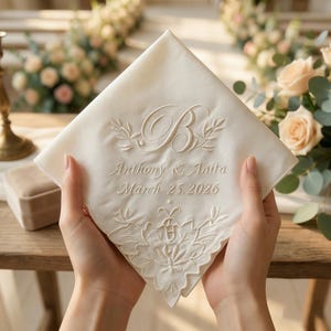 Pode incluir: Lenço de cor marfim com bordados florais e de folhas. O lenço apresenta a letra "B", os nomes "Anthony & Anita" e a data "25 de março de 2026".