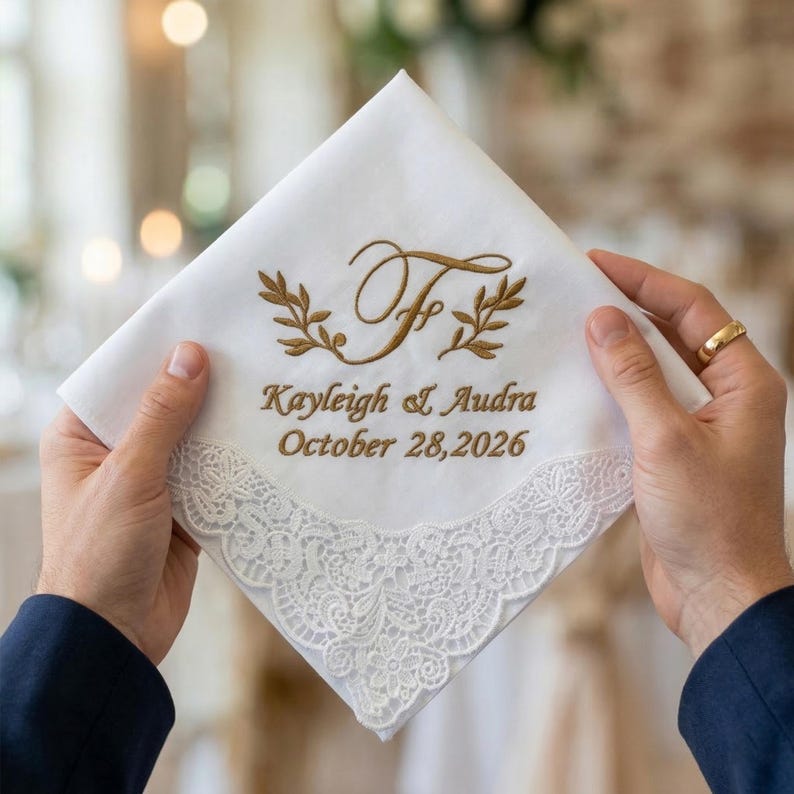Peut inclure: Mouchoir blanc avec broderie dor&eacute;e et bordure en dentelle. Le mouchoir porte les noms "Kayleigh & Audra" et la date "28 octobre 2026", ainsi qu'une initiale d&eacute;corative et des feuilles de laurier.