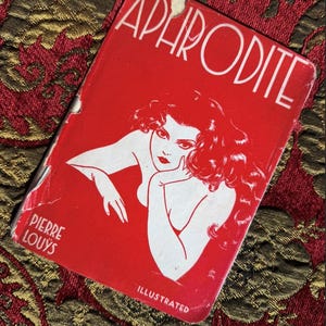 Puede incluir: Una portada de libro vintage roja con el título "APHRODITE" en blanco. Presenta una ilustración blanca de una mujer, el nombre del autor Pierre Louÿs y la palabra "ILLUSTRATED".