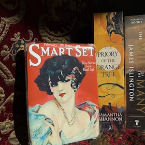 Puede incluir: Una colección de libros, incluyendo una portada de la revista "Smart Set" con una ilustración vintage de una mujer. La portada de la revista es roja con el texto "Smart Set" y "February - 25 Cents". Otros libros incluyen "The Priory of the Orange Tree" y "The Will of the Many".