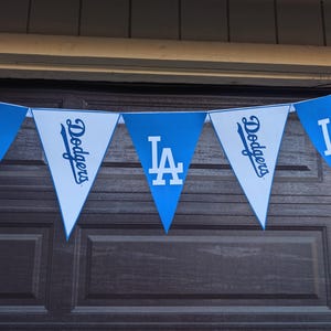 May include: A pennant banner featuring blue and white triangular flags. The flags display the Los Angeles Dodgers logo and team name in white lettering. The banner is hanging against a brown background.