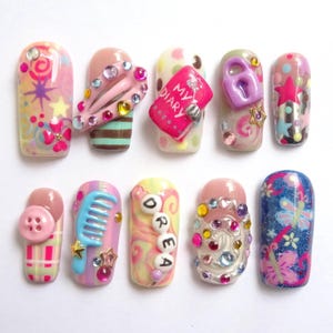 May include: A collection of ten decorated artificial nails. Each nail features a unique design, including rhinestones, charms, and painted patterns. Some nails have text that reads "MY DIARY" and "DREAM". Colors include pink, blue, yellow, and white.