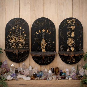 Può includere: Tre mensole da parete in legno nero con la parte superiore ad arco, ciascuna con disegni celesti dorati. Le mensole hanno una base semicircolare e sono decorate con una farfalla, una mano e fasi lunari. Cristalli e piante sottostanti.