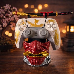 May include: A white and gray PlayStation 5 controller with gold accents and a samurai-themed design rests on a red Oni mask stand. A miniature katana sits above the controller. The stand is silver and the background is blurred.