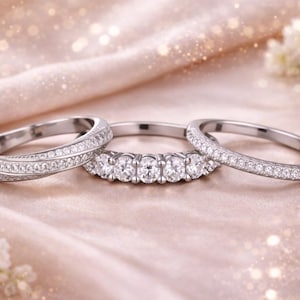 May include: Three silver rings with sparkling white gemstones. One ring features a row of five larger stones, while the other two are adorned with smaller stones. The rings are displayed on a soft, pink fabric.