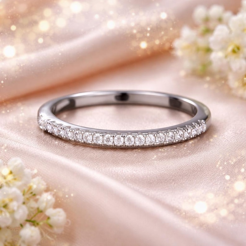 May include: A silver band ring with a row of small, clear gemstones set along the top. The ring is displayed on a soft, pink fabric, with white flowers in the background. The ring is a delicate piece of jewelry.
