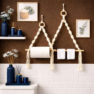 May include: A macrame towel rack with a wooden dowel and two white towels. The rack is suspended from a wooden ring. The background features a brown wall, white tile, and decorative items, including vases and framed artwork.