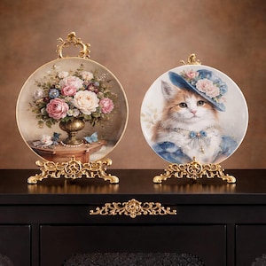 Puede incluir: Dos platos decorativos sobre soportes dorados ornamentados. Uno presenta un arreglo floral en un jarrón dorado. El otro representa un gato con un sombrero azul con rosas rosas. Ambos platos tienen bordes dorados.