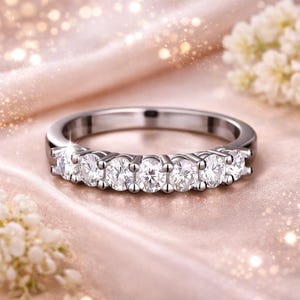 May include: A silver band ring with seven round, clear gemstones set in a row. The ring is displayed on a soft, pink surface with blurred floral accents, creating a delicate and elegant aesthetic. The ring is a piece of jewelry.