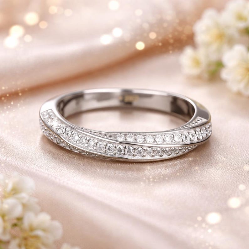 May include: A silver ring with a curved design, encrusted with small, clear gemstones. The ring is set against a soft, peach-colored background with blurred lights and flowers, creating a delicate and elegant aesthetic.