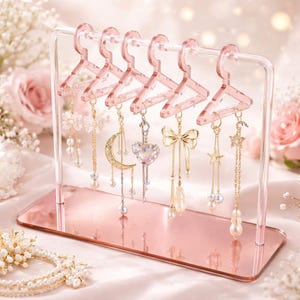 Op de afbeelding: Een roze acryl oorbellen displaystandaard met zes miniatuur hanger-vormige houders. De standaard bevat diverse bungelende oorbellen met parel-, ster-, hart- en strikdesigns. De standaard staat op een roze basis.