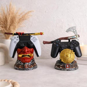 May include: Two gaming controller stands with decorative elements. One has a white controller with a red Oni mask base and a katana. The other has a black controller with a gold shield base and an axe. Both stands feature metallic detailing.