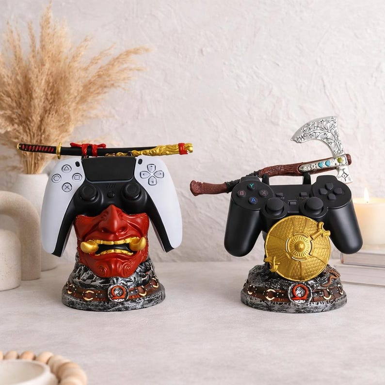 May include: Two decorative gaming controller stands. One features a white controller with a red and gold Oni mask base and a katana. The other has a black controller with a gold shield base and an axe. Both stands are detailed with metallic accents.