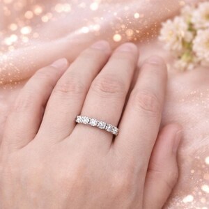 May include: A silver ring with five round, clear gemstones set in a row. The ring is on a person's finger, set against a soft pink background with bokeh lights and white flowers.