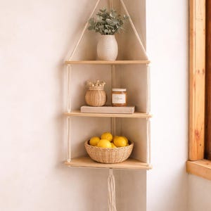 Op de afbeelding: Een hoekplank met drie niveaus van licht hout, opgehangen aan een crèmekleurig touw. De bovenste plank bevat een witte vaas met groen. De middelste plank heeft een klein gevlochten mandje en een pot. De onderste plank heeft een gevlochten mand met citroenen.
