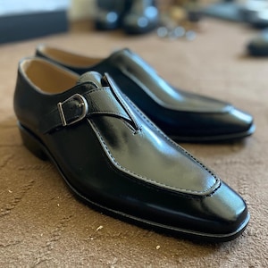May include: A pair of black leather dress shoes with a buckle closure. The shoes have a sleek, polished appearance with a pointed toe and a stitched design along the edges. The shoes are displayed on a brown carpet.
