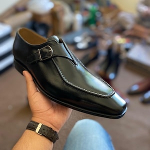 May include: Black leather dress shoe with a buckle closure. The shoe features a square toe design and white stitching along the edges. The shoe is held in a person's hand, with a watch visible on the wrist. Other shoes are visible in the background.