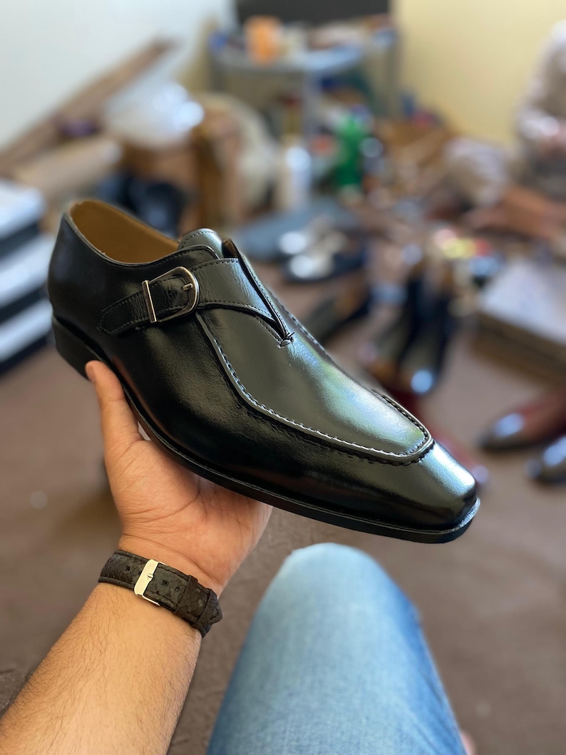 May include: Black leather dress shoe with a buckle closure. The shoe features a square toe design and white stitching along the edges. The shoe is held in a person's hand, with a watch visible on the wrist. Other shoes are visible in the background.