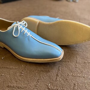 Pu&ograve; includere: Scarpe in pelle azzurra con lacci e cuciture bianche. Le scarpe hanno la punta arrotondata e una suola colorata chiara. Una scarpa &egrave; a fuoco, mentre l'altra &egrave; leggermente sfocata sullo sfondo. Le scarpe sono su una superficie marrone.