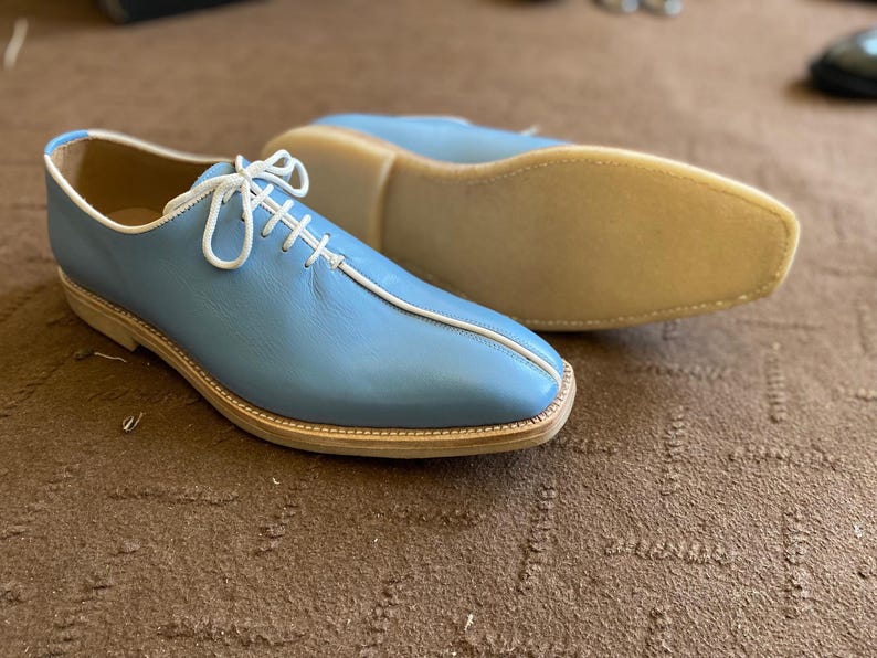 Pu&ograve; includere: Scarpe in pelle azzurra con lacci e cuciture bianche. Le scarpe hanno la punta arrotondata e una suola colorata chiara. Una scarpa &egrave; a fuoco, mentre l'altra &egrave; leggermente sfocata sullo sfondo. Le scarpe sono su una superficie marrone.