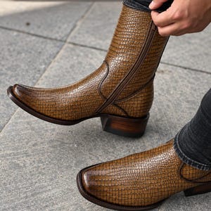 Peut inclure: Une paire de bottes en cuir marron avec un motif texturé en peau de crocodile, un bout pointu et une fermeture éclair latérale. Les bottes présentent un talon en bois empilé et une semelle marron foncé, avec une finition polie et dégradée.