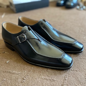 May include: A pair of black leather dress shoes with a buckle closure. The shoes have a polished finish and a stitched design around the toe. The shoes are displayed on a brown surface.