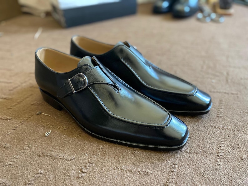 May include: A pair of black leather dress shoes with a buckle closure. The shoes have a polished finish and a stitched design around the toe. The shoes are displayed on a brown surface.