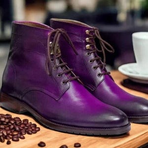 Può includere: Un paio di stivali in pelle viola intenso con lacci marroni. Gli stivali sono su una superficie di legno, accanto a chicchi di caffè e una tazza e piattino bianchi. Calzature eleganti.