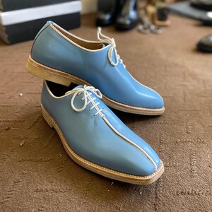 Pu&ograve; includere: Un paio di scarpe eleganti in pelle blu chiaro con lacci e finiture bianche. Le scarpe hanno un design a punta quadrata e una suola colorata chiara. Le scarpe sono esposte su una superficie marrone.