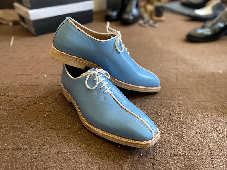 Pu&ograve; includere: Un paio di scarpe eleganti in pelle blu chiaro con lacci e finiture bianche. Le scarpe hanno un design a punta quadrata e una suola colorata chiara. Le scarpe sono esposte su una superficie marrone.