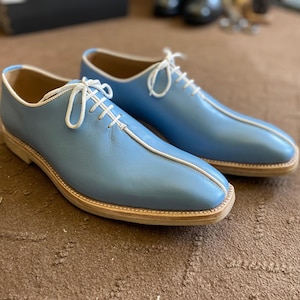 Pu&ograve; includere: Un paio di scarpe eleganti in pelle azzurra con lacci e finiture bianche. Le scarpe hanno un design classico con punta arrotondata e suola chiara. Sono esposte su una superficie marrone.