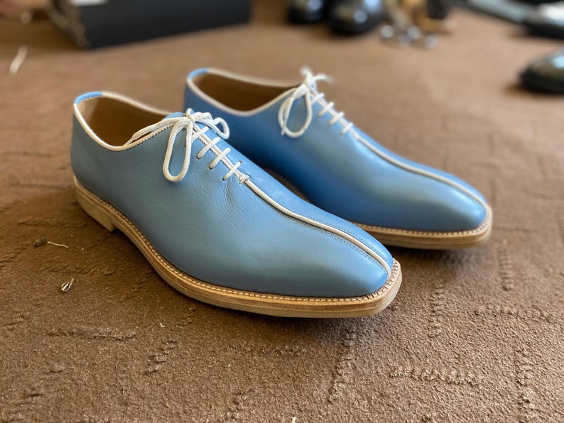 Pu&ograve; includere: Un paio di scarpe eleganti in pelle azzurra con lacci e finiture bianche. Le scarpe hanno un design classico con punta arrotondata e suola chiara. Sono esposte su una superficie marrone.