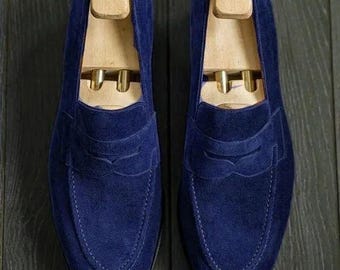May include: A pair of navy blue suede loafers with wooden shoe trees inserted. The shoes have a classic penny loafer design with a stitched vamp and a rounded toe. The background is a dark wood surface.