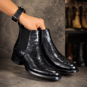 Puede incluir: Un par de botas Chelsea negras con un acabado de cuero texturizado estilo avestruz y un patrón de puntos distintivo. Las botas tienen paneles laterales elásticos, punta redondeada, un tacón bajo y una lengüeta en la parte posterior.