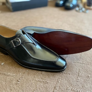 May include: Black leather dress shoes with a buckle closure. The shoes have a polished finish and a classic design. One shoe is upright, while the other is lying on its side, revealing the wooden sole. A timeless footwear choice.