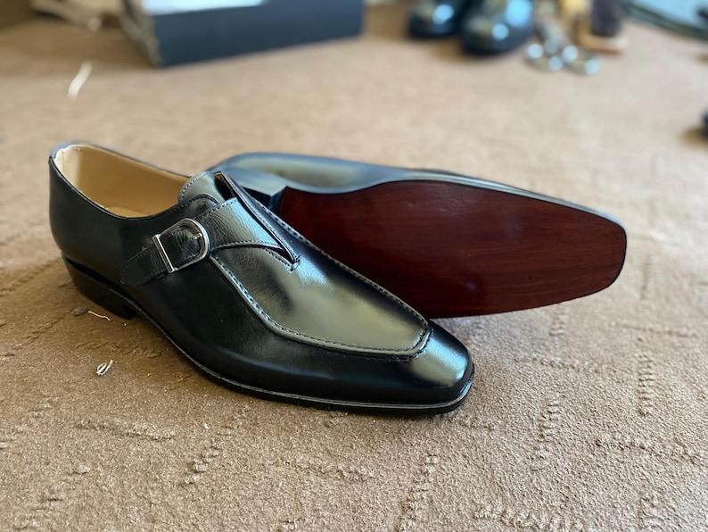 May include: Black leather dress shoes with a buckle closure. The shoes have a polished finish and a classic design. One shoe is upright, while the other is lying on its side, revealing the wooden sole. A timeless footwear choice.