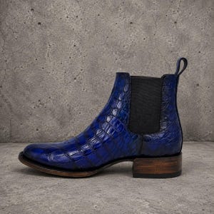 Op de afbeelding: Een blauwe Chelsea boot met een textuur van krokodillenleer op het bovenwerk. De laars is voorzien van een zwart elastisch zijpaneel, een treklipje bij de hiel en een gestapelde houten hak met een donkerbruine afwerking aan de onderkant.