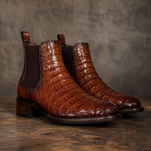 Puede incluir: Un par de botas Chelsea marrones con un acabado de cuero texturizado con relieve de cocodrilo, paneles laterales elásticos de color marrón oscuro, una trabilla en el talón, un tacón de madera apilado, suela oscura y punta redondeada.