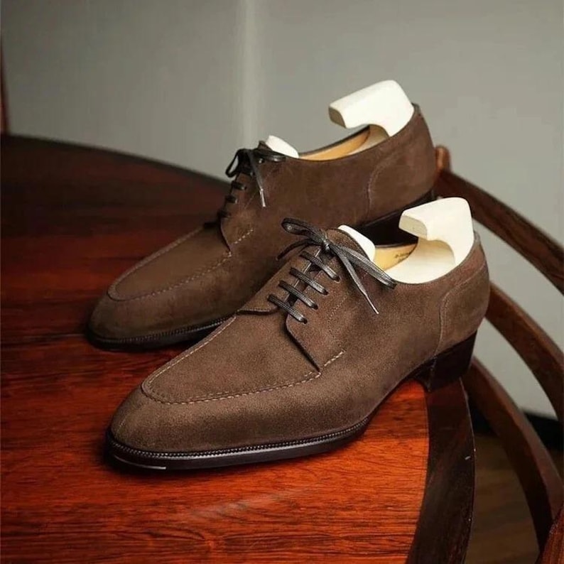 May include: A pair of brown suede dress shoes with black laces. The shoes have a classic design with a rounded toe and a low heel. Shoe trees are inserted in the shoes. The shoes are displayed on a wooden surface.
