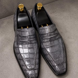 Może przedstawiać: Para szarych mokasynów o fakturze skóry krokodyla. Buty posiadają klasyczny pasek typu penny loafer na przyszwie oraz czarne prawidła z polerowanymi, złotymi metalowymi gałkami umieszczonymi na górze każdego z nich.
