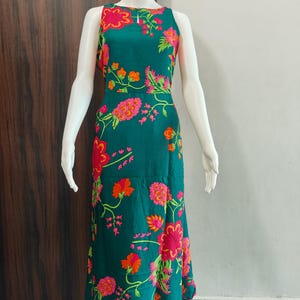 Op de afbeelding: Een mouwloze, enkellange jurk in een diepe groenblauwe kleur met een levendig bloemenpatroon in tinten felroze, oranje en lichtgroen. De jurk heeft een ronde halslijn met een klein sleutelgat-detail in het midden.