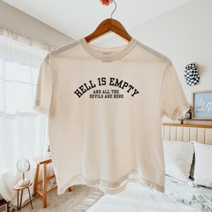 May include: A cream-coloured short-sleeve t-shirt featuring black text printed on the chest. The text reads "HELL IS EMPTY" in an arched font, followed by "AND ALL THE DEVILS ARE HERE" in a straight line below it. The shirt has a crew neckline.